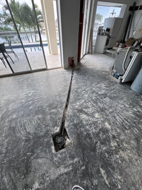 Plumbing access cut into a concrete slab floor leading to a shutoff valve, with a screened pool lanai visible through the sliding door
